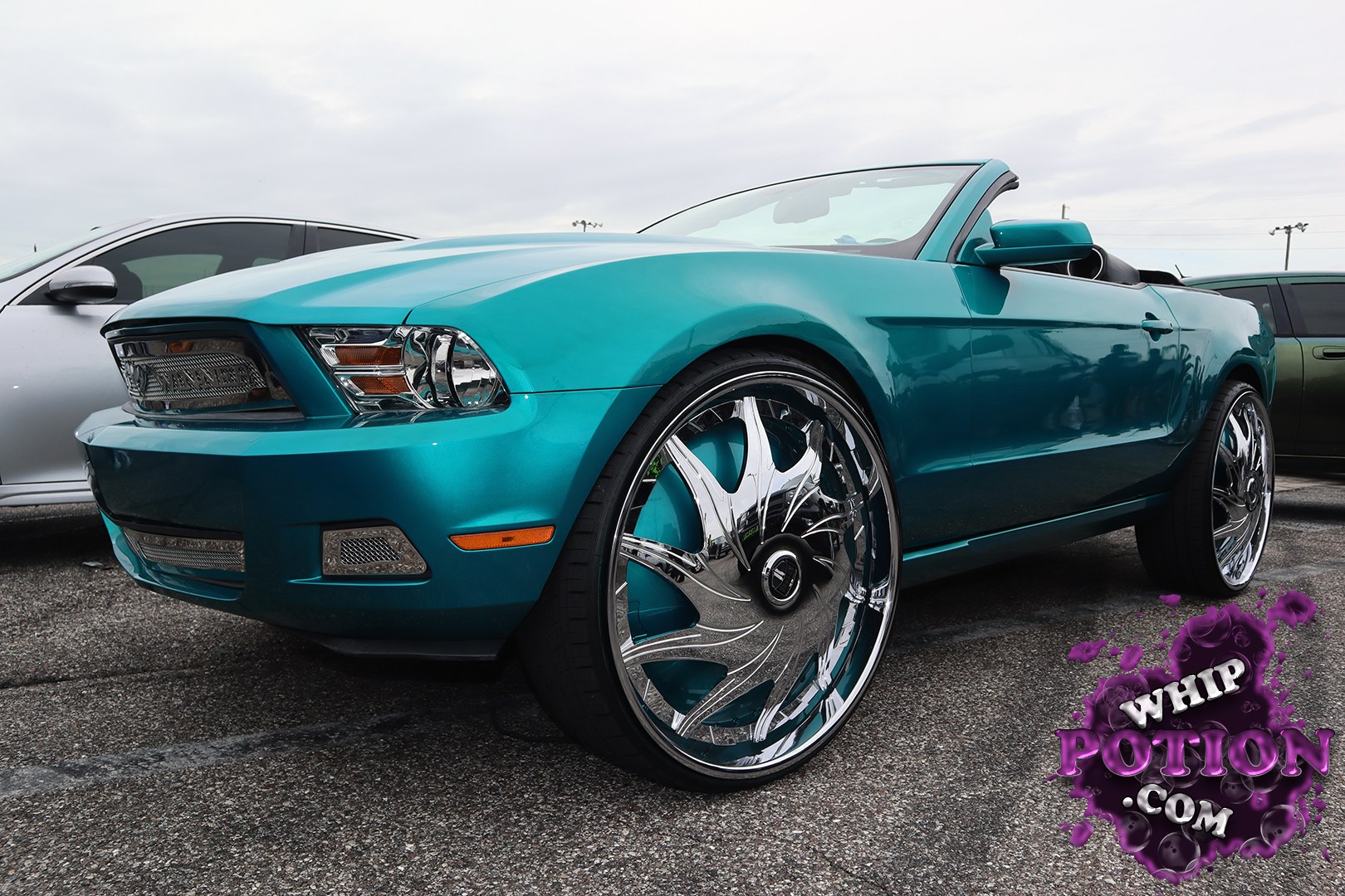 A teal Ford Mustang convertible sits on massive chrome wheels that fill the arches with a serious custom presence. The clean body color, tinted lights, and low-slung stance give it a polished show-car look, while the open top and oversized rims bring plenty of street flair without going overboard.