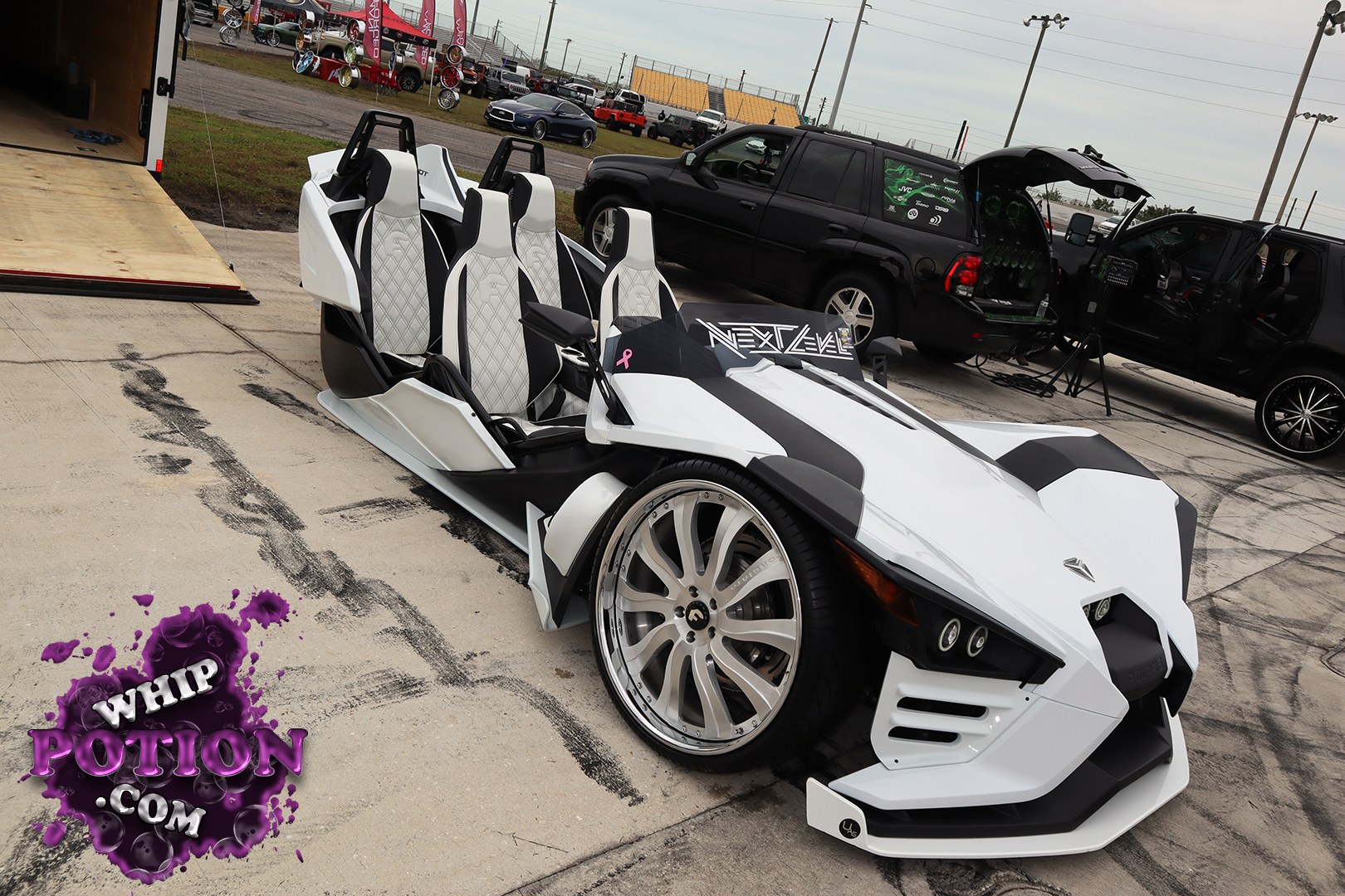 A wild open-air custom three-wheeler with sharp wedge bodywork, dual bucket seats, and a white-and-black finish that really leans into the futuristic vibe. The oversized billet-style wheels tuck tight under the low nose, while the exposed cabin and sculpted panels give it serious show-floor presence without losing that homemade hot-rod attitude.