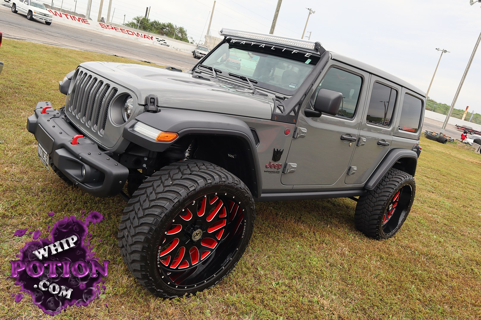 This Jeep Wrangler Unlimited has a tough, purposeful look with a satin-gray finish, blackout trim, and a tall stance on massive forged wheels wrapped in aggressive off-road rubber. The light bar, hood-mounted accents, and heavy-duty front bumper give it a clean overland-meets-show-truck vibe, while the red wheel detailing adds just enough flash to keep it interesting.
