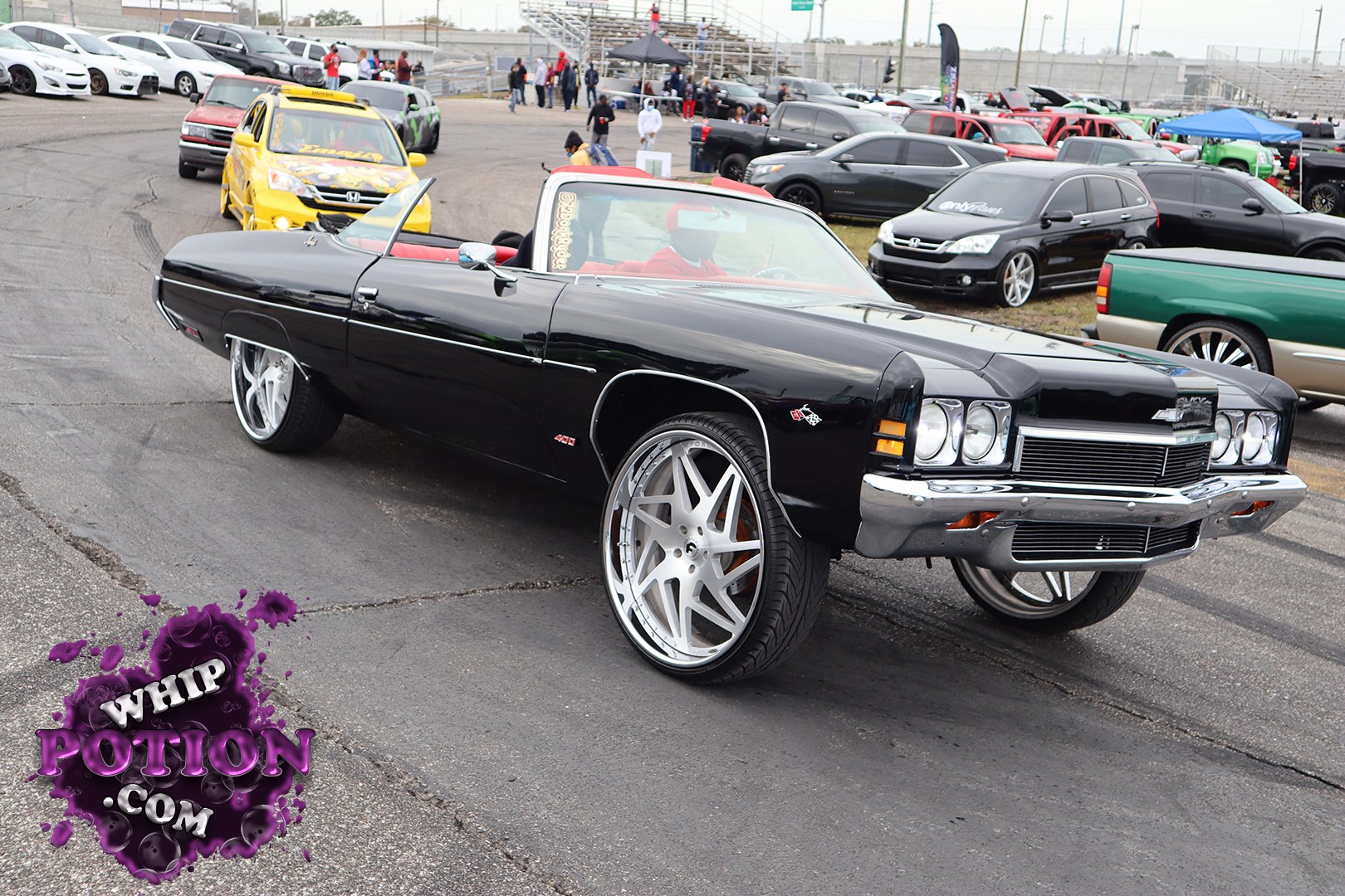 A classic Chevrolet Impala convertible gets a bold custom treatment here, finished in deep gloss black with a red interior and massive polished multi-spoke wheels tucked under the fenders. The long nose, stacked headlights, chrome-heavy front bumper, and clean lowered stance give it serious street presence without losing that old-school Chevy swagger.