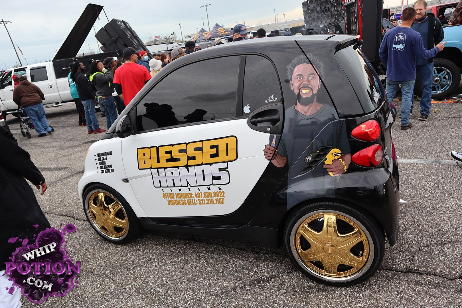 A Smart Fortwo gets full custom treatment here, with a two-tone black-and-white wrap, oversized gold billet-style wheels, and a bold “Blessed Hands” graphics package running down the doors. The cartoon portrait on the rear quarter adds a personal touch, while the tiny footprint and slammed stance give the little car serious show-floor attitude.