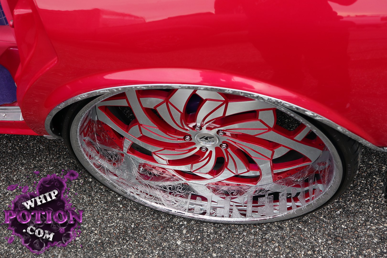 A deep red customized sedan rides on a massive engraved billet wheel with a bright red-and-silver swirl design and “BEN ETRU” cut into the lip. The tucked tire and polished trim give it that tight, show-ready stance, and the attention to detail in the wheel alone says this build was made for close inspection.