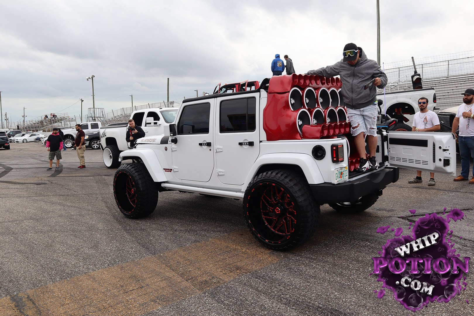 This Jeep Wrangler Unlimited is built to make noise and look good doing it, with a clean white body, oversized black-and-red wheels, and a huge custom rear audio wall packed with subwoofers. The red speaker enclosure and open tailgate give it a full show-truck vibe, while the lifted stance and knobby tires keep it rooted in off-road culture.