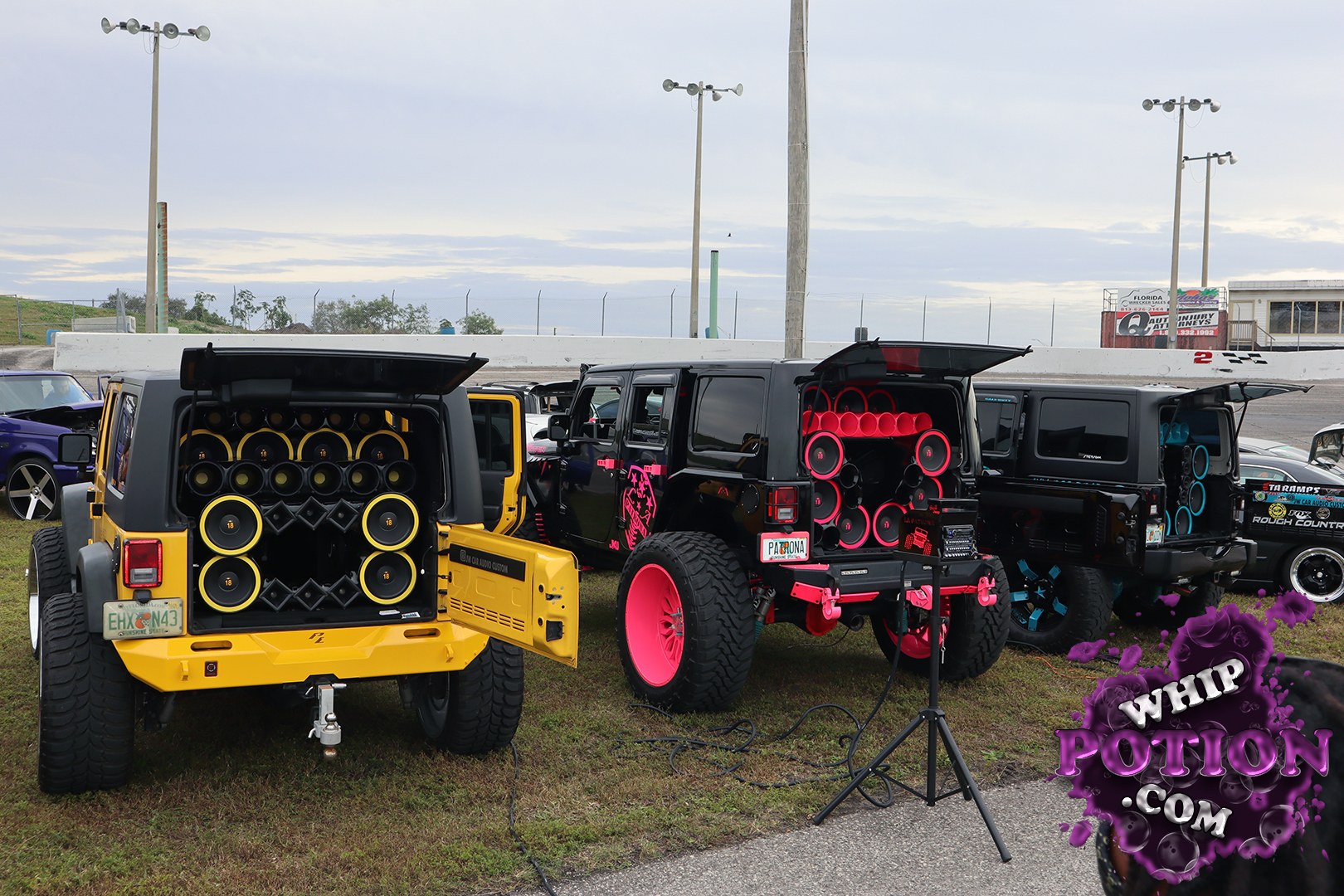 A row of Jeep Wrangler builds gets wild with full custom audio trunks and bold color-matched accents. The yellow two-door brings a wall of yellow-ringed speakers, while the black rigs lean into neon pink and blue details, deep-dish wheels, and chunky tires. It’s all about big sound, big personality, and serious trail-ready attitude.