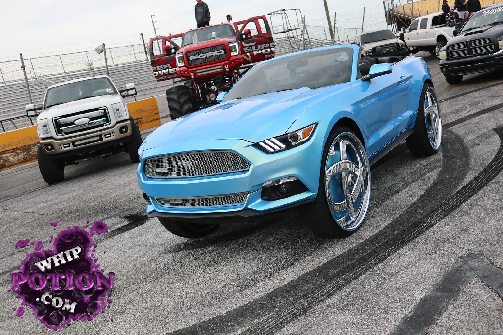 A bright blue Ford Mustang convertible sets the tone with its clean mesh grille, triple-bar LED headlights, and oversized chrome blades that give it a show-car attitude. The stance is dialed in low enough to look deliberate without going overboard, and the open-top setup keeps the cockpit in full view for that polished, street-ready feel.
