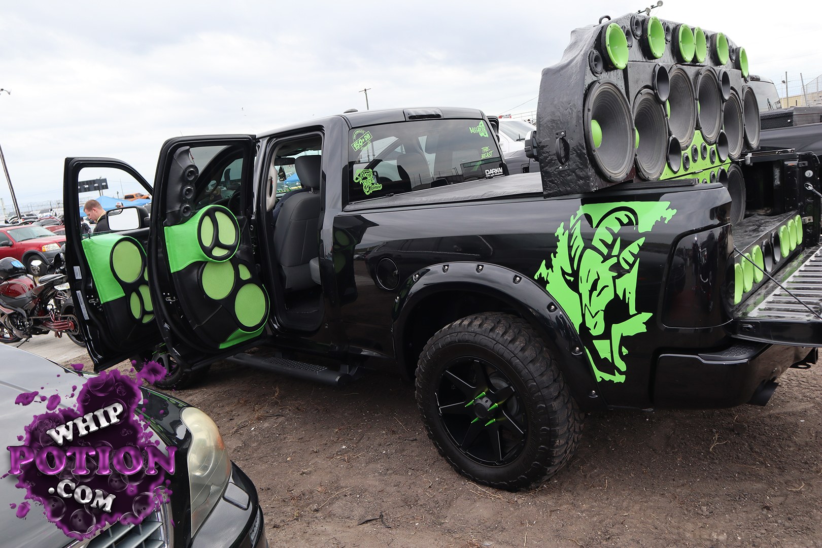 A black crew-cab pickup turns up the attitude with neon green graphics, matching speaker-panel door cards, and a massive bed-mounted wall of audio gear. The stance sits on chunky off-road tires and black wheels, while the open rear and custom trim make it clear this build is all about loud visuals and even louder bass.
