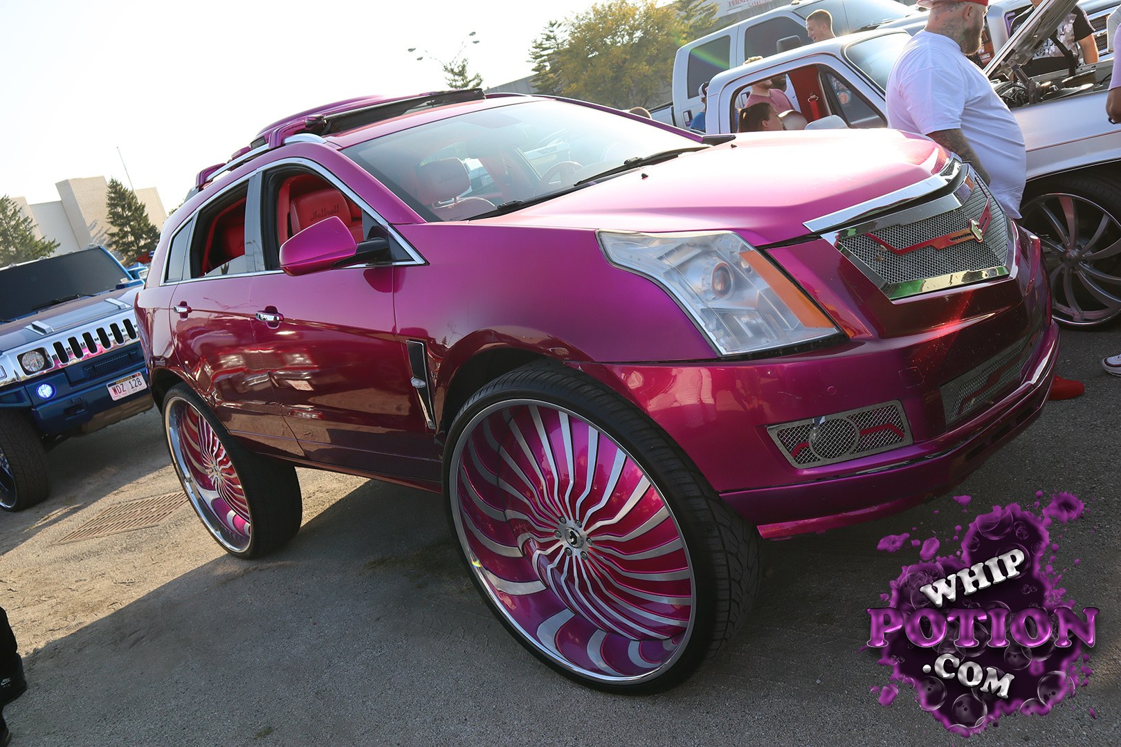 This magenta Cadillac SRX leans hard into custom culture with oversized 30s-style wheels, a matching paint-matched grille, and a slammed-but-not-overdone stance. The red interior pops through the open windows, and the whole build has that polished show-truck feel without losing the factory SUV lines.