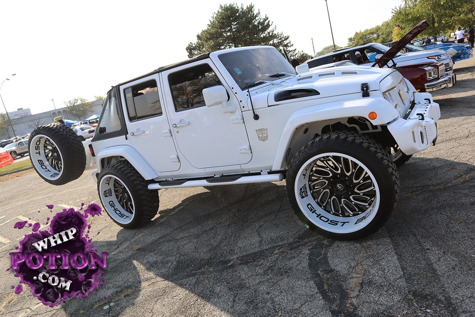 A white Jeep Wrangler Unlimited gets a full custom treatment with a lifted stance, chunky off-road tires, and big two-tone Ghost rims that really set it off. The clean black soft top, tube-style side steps, and hood vents give it a purposeful, trail-ready look, while the spare matching wheel out back ties the build together nicely.
