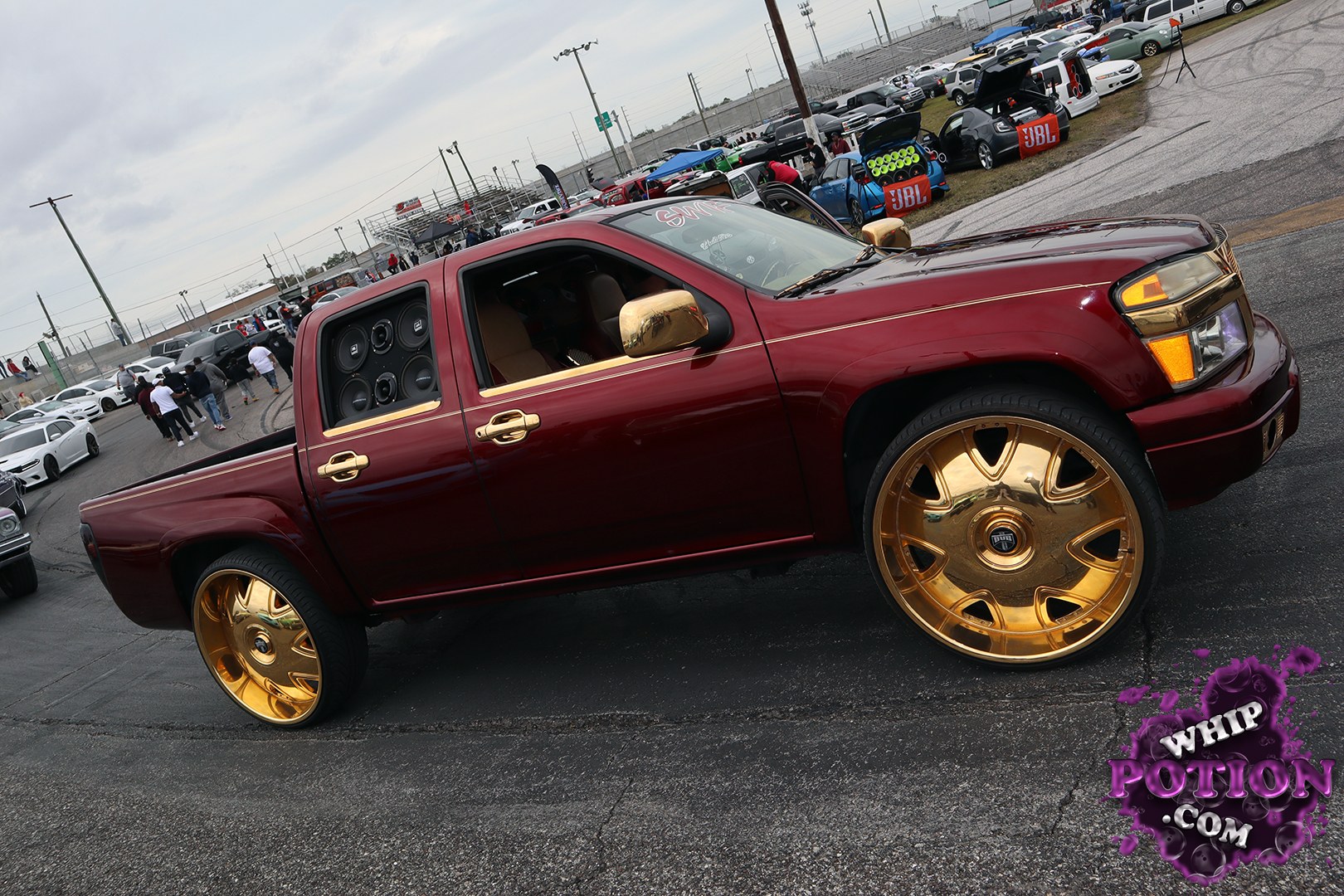A slammed Chevrolet Silverado crew cab brings serious style with deep burgundy paint, gold mirror caps, and massive matching wheels that fill the wells nicely. The rear door panel is opened up to show a clean multi-speaker audio setup, while the tidy stance and smooth bodywork give this truck a polished, show-ready presence.