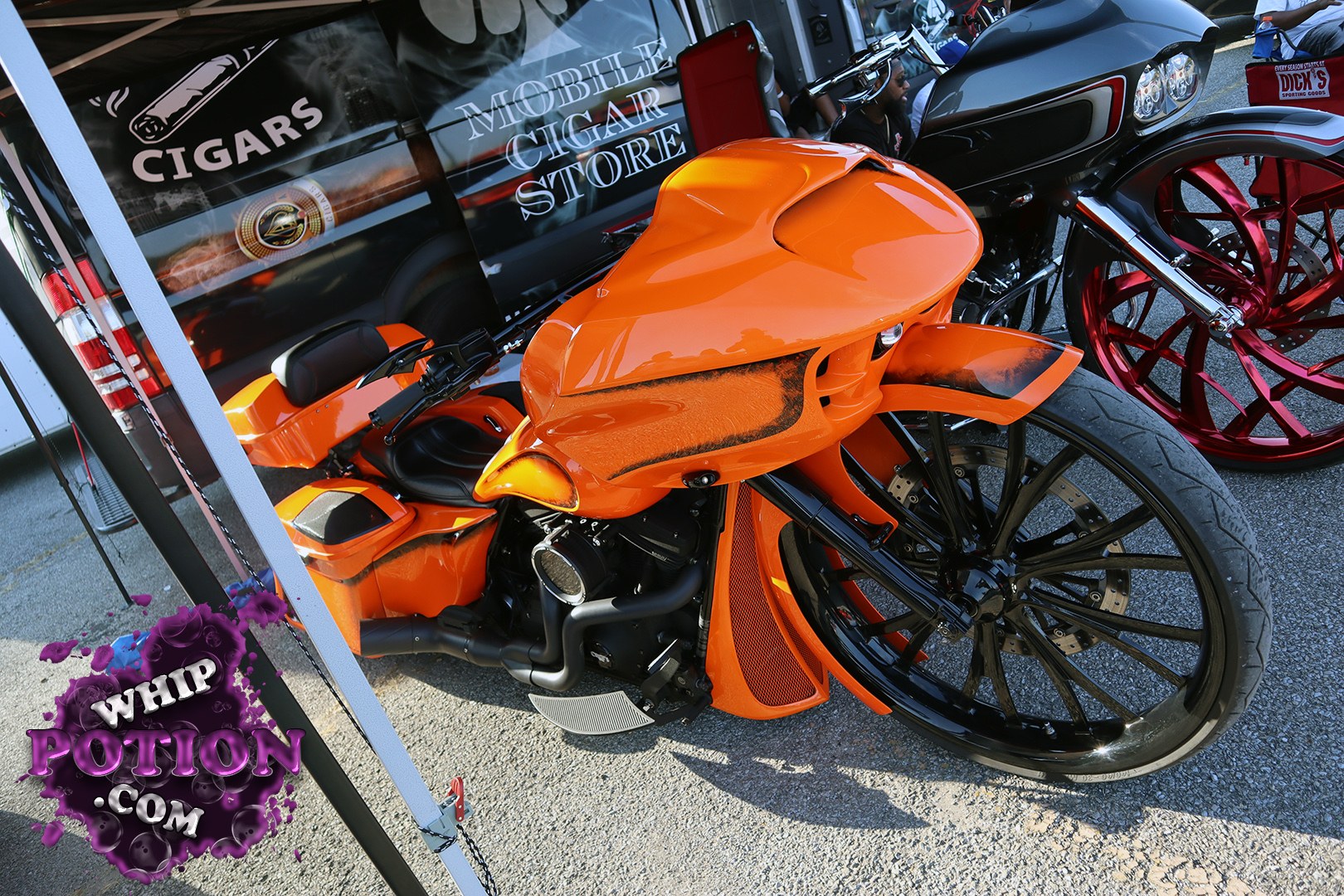 This orange custom bagger has serious show-bike presence, with a slammed stance, stretched front end, and massive black-and-chrome wheel tucked under that sculpted fairing. The bodywork is smooth and tightly finished, and the deep gloss paint pops hard against the black mechanicals. Behind it, the red-spoke tourer adds even more custom flavor to the lineup.