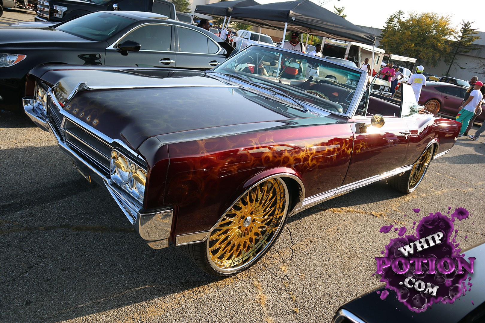 A clean mid-’60s Oldsmobile Cutlass convertible sits low on oversized gold billets, with deep burgundy paint, a black top, and subtle flame graphics licking across the front fender. The chrome nose, low stance, and shaved-looking details give it that crisp pro-street-meets-cruise-night vibe that car people always stop to study.