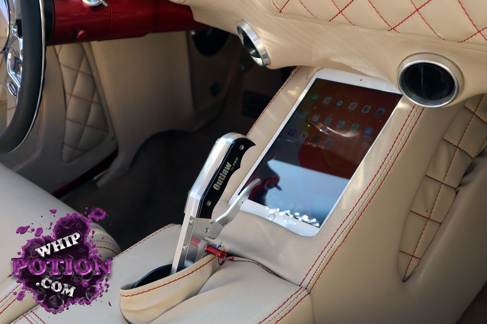 A meticulously finished custom interior wraps the cabin in cream leather with red diamond stitching, accented by a deep red steering column and polished billet touches. The standout is a tablet-mounted center console setup with a sculpted shifter and cupholders, giving the build a modern street-cruiser vibe without losing its handcrafted feel.