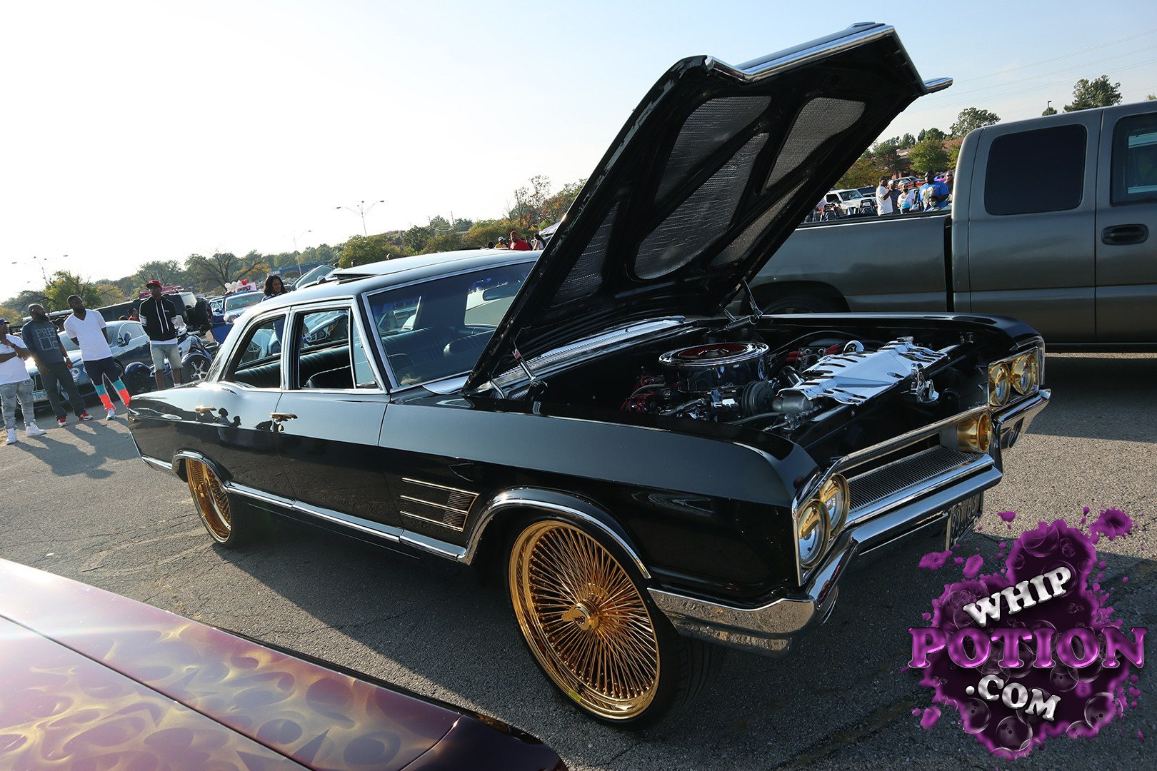 A black classic Chevrolet Impala wears a clean resto-mod look with a gloss body, chrome trim, and gold wire wheels that set it off perfectly. The hood is propped to show off a polished V8 bay, while the low stance and tucked tires give it that tasteful custom street-cruiser attitude.