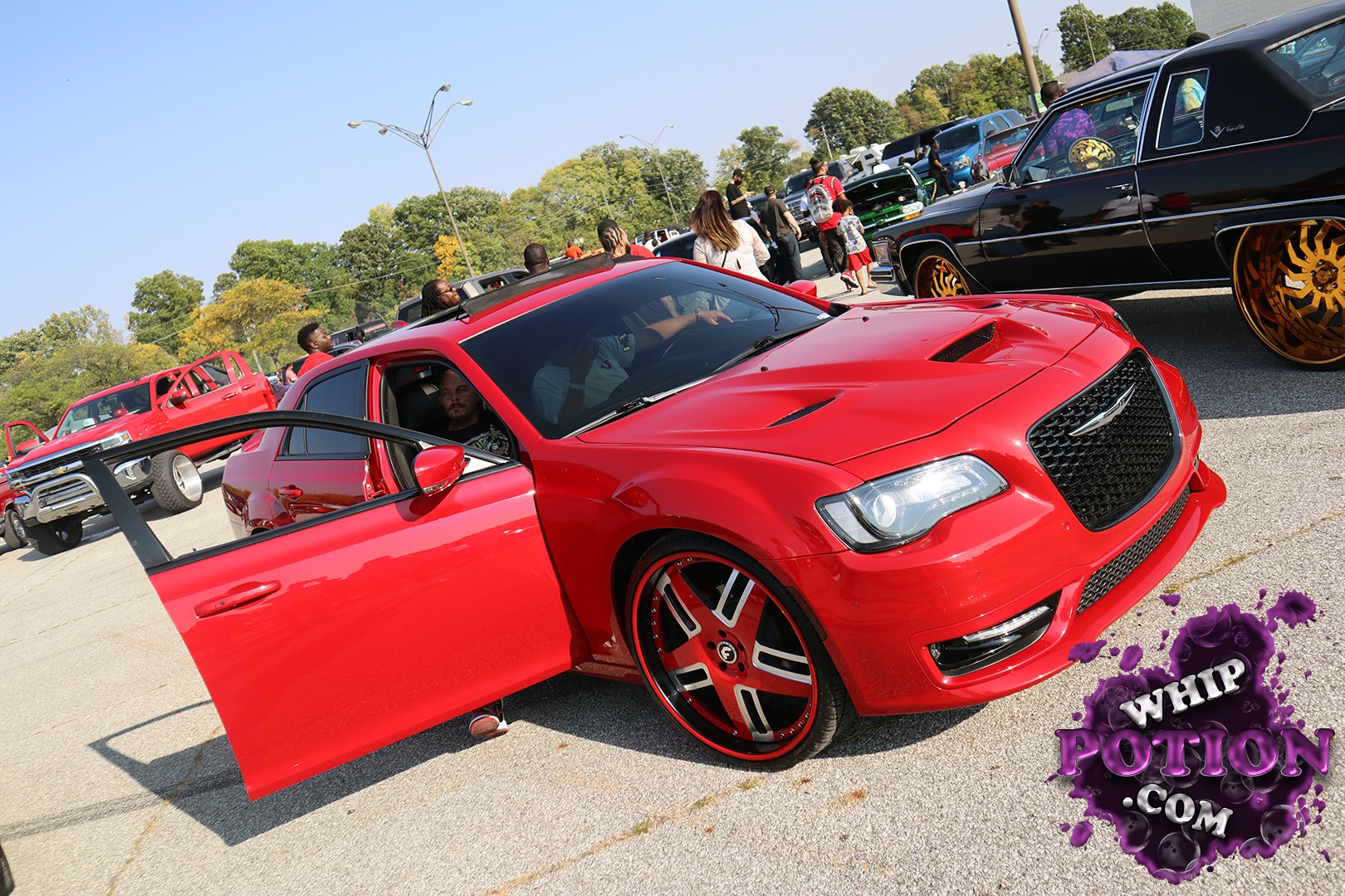 A red Chrysler 300 gets a heavy custom treatment with a smooth grille, vented hood, and oversized billet-style wheels wrapped in thin rubber. The blacked-out trim and dramatic door setup give it a clean, show-ready presence, while the lowered stance and color-matched details lean hard into modern street-style luxury.