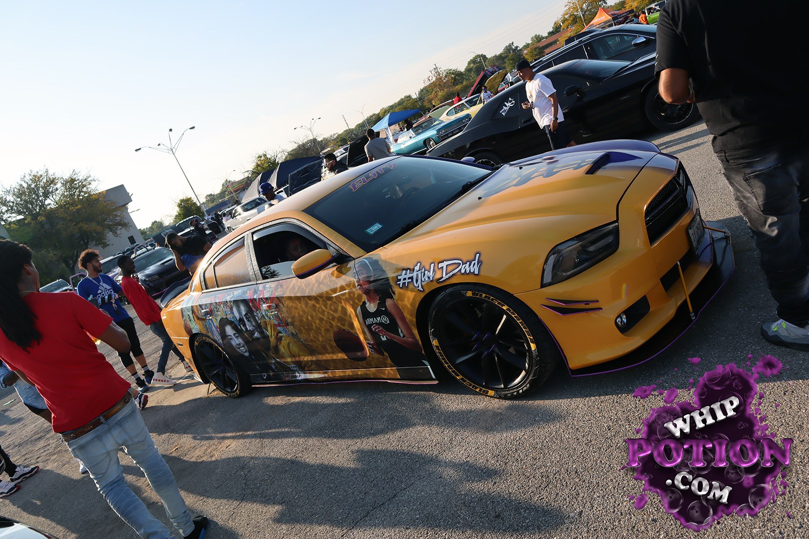 A slammed orange Dodge Charger wears a full custom graphic wrap with celebrity portraits and bold “#fwm Dad” lettering, plus deep-dish black wheels with polished lips and purple accent trim. The body sits low and clean on the pavement, with a sharp front splitter and tinted glass giving it that serious street build look.