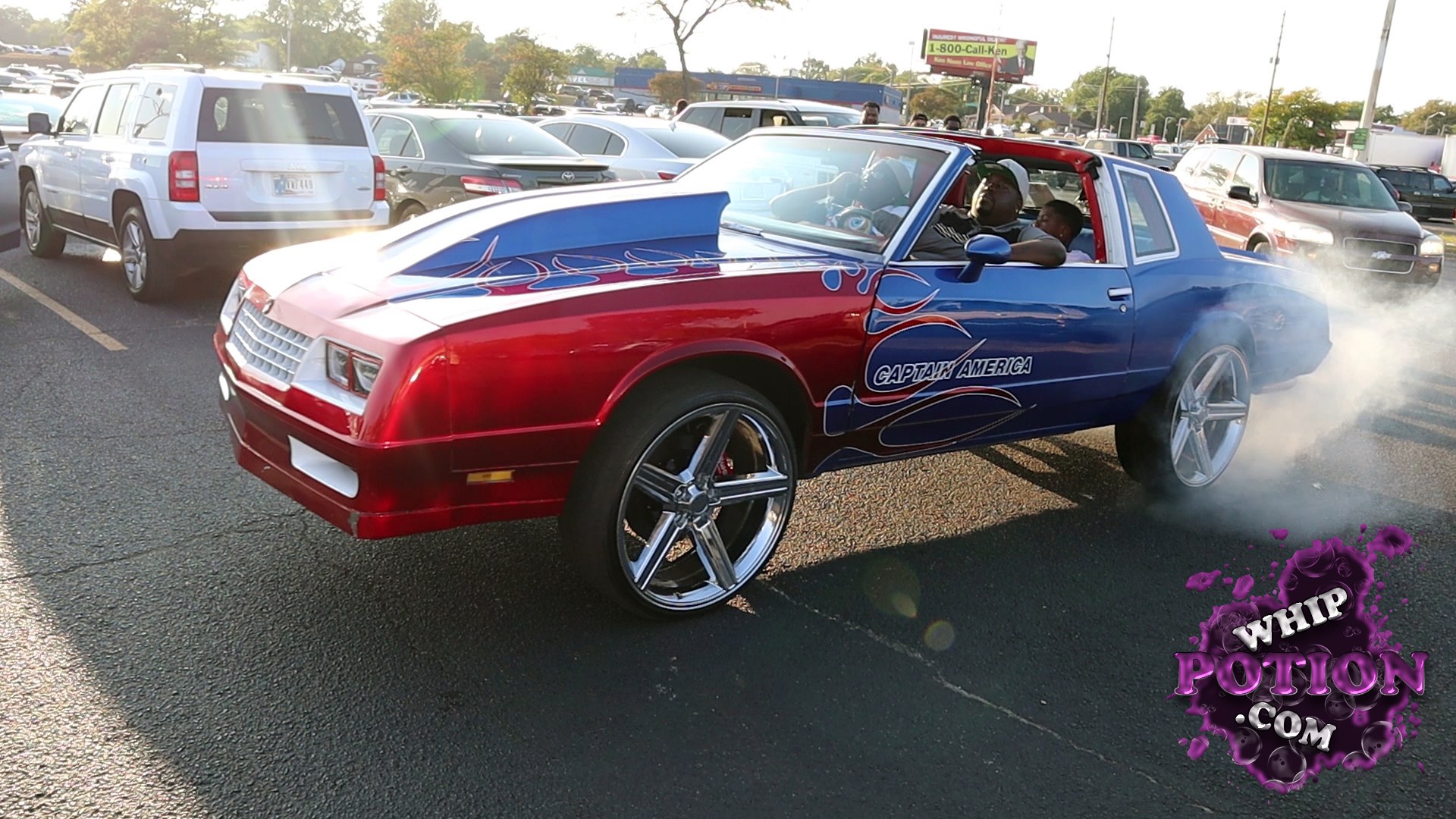 A wild two-tone Chevrolet Monte Carlo wears a deep red-and-blue custom paint job with flame graphics and “Captain America” script down the side. The big chrome five-spokes and tall stance give it that boulevard-cruiser look, while the open interior and bold styling make it feel built to be seen, not just driven.