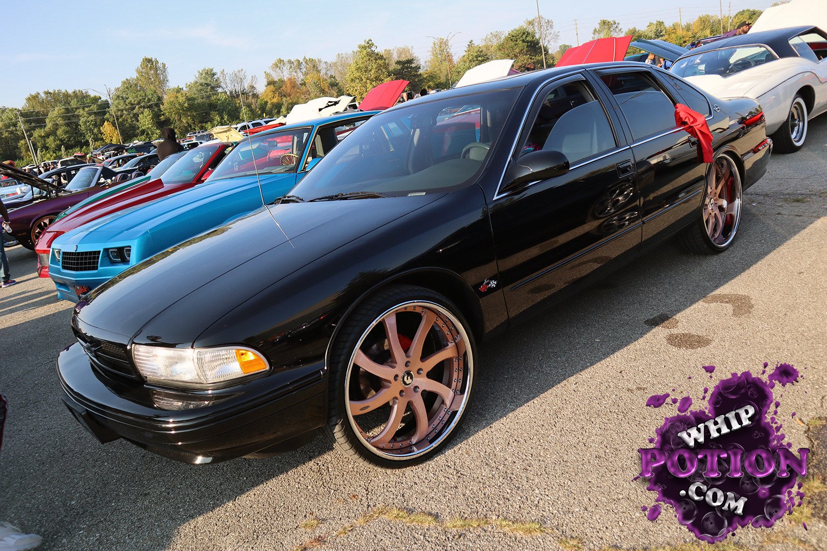 A clean black Chevrolet Impala sedan sits low on oversized bronze billets, with a glossy finish and darkened glass that give it a sharp, street-ready look. The mirror-like bodywork and red ribbon on the rear door add a little show flair, while the stance and wheel choice give this build plenty of presence without going overboard.